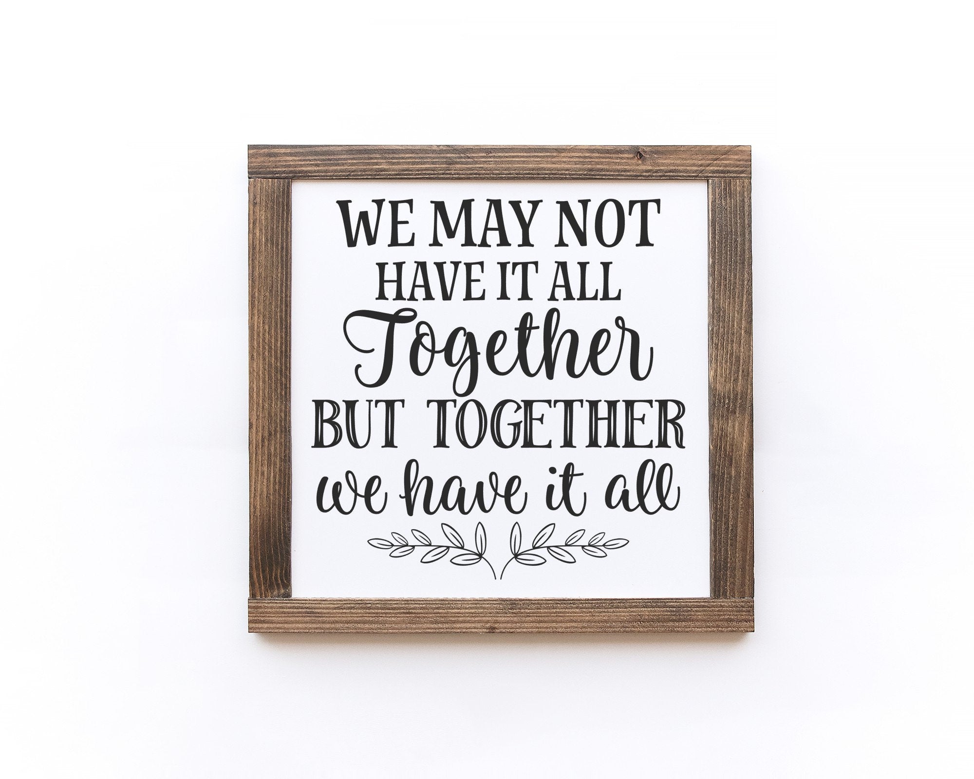 A handmade wooden sign with a white background and black lettering that reads 'We May Not Have It All Together But Together We Have It All'.