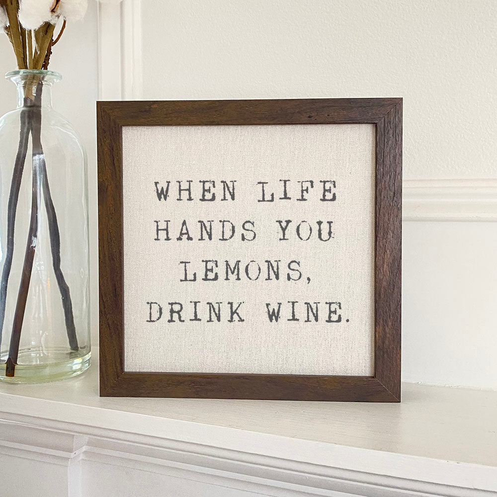 Framed sign with the phrase 'When Life Hands You Lemons, Drink Wine' in a stylish wood frame, displayed on a shelf.