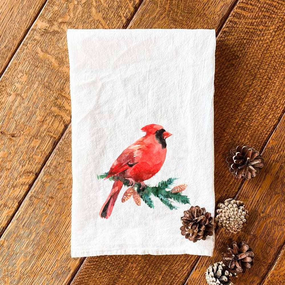 A cotton tea towel featuring a vibrant cardinal perched on an evergreen branch, showcasing its beautiful design and hemmed edges.