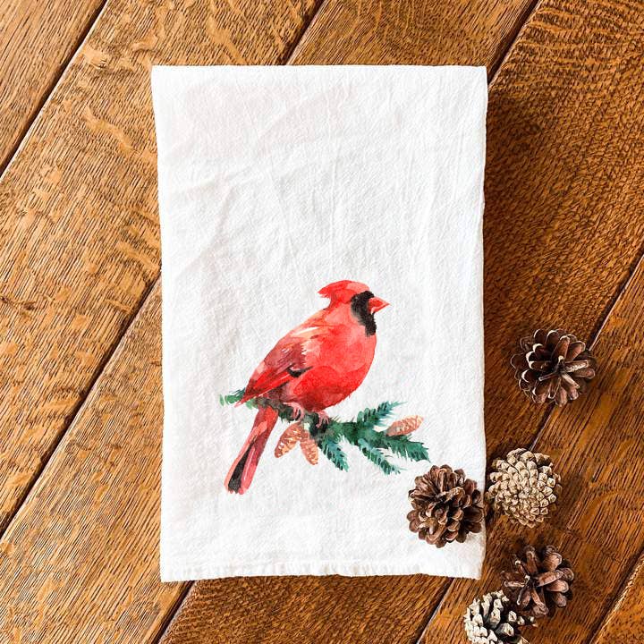 A cotton tea towel featuring a vibrant cardinal perched on an evergreen branch, showcasing its beautiful design and hemmed edges.