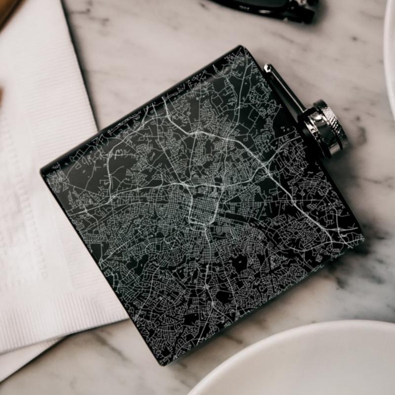 Matte black hip flask featuring a custom engraved map of Charlotte, North Carolina with coordinates, accompanied by a canvas bag and funnel.