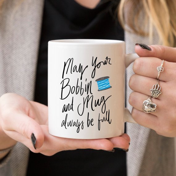 A high-quality ceramic coffee mug featuring the humorous phrase 'May Your Bobbin And Mug Always Be Full', perfect for sewing enthusiasts.