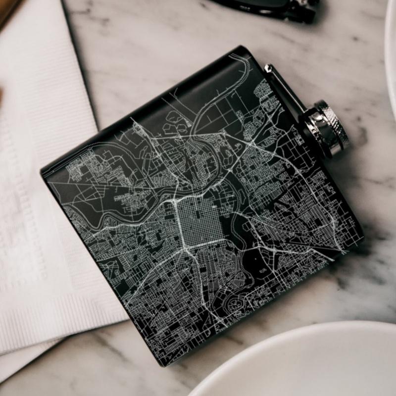 Matte black hip flask featuring a custom engraved map of Sacramento, California with coordinates, accompanied by a canvas bag and funnel.