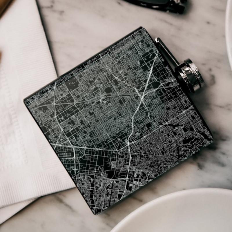 Matte black hip flask featuring a custom engraved map of Santa Ana, California with coordinates, accompanied by a canvas bag and funnel.