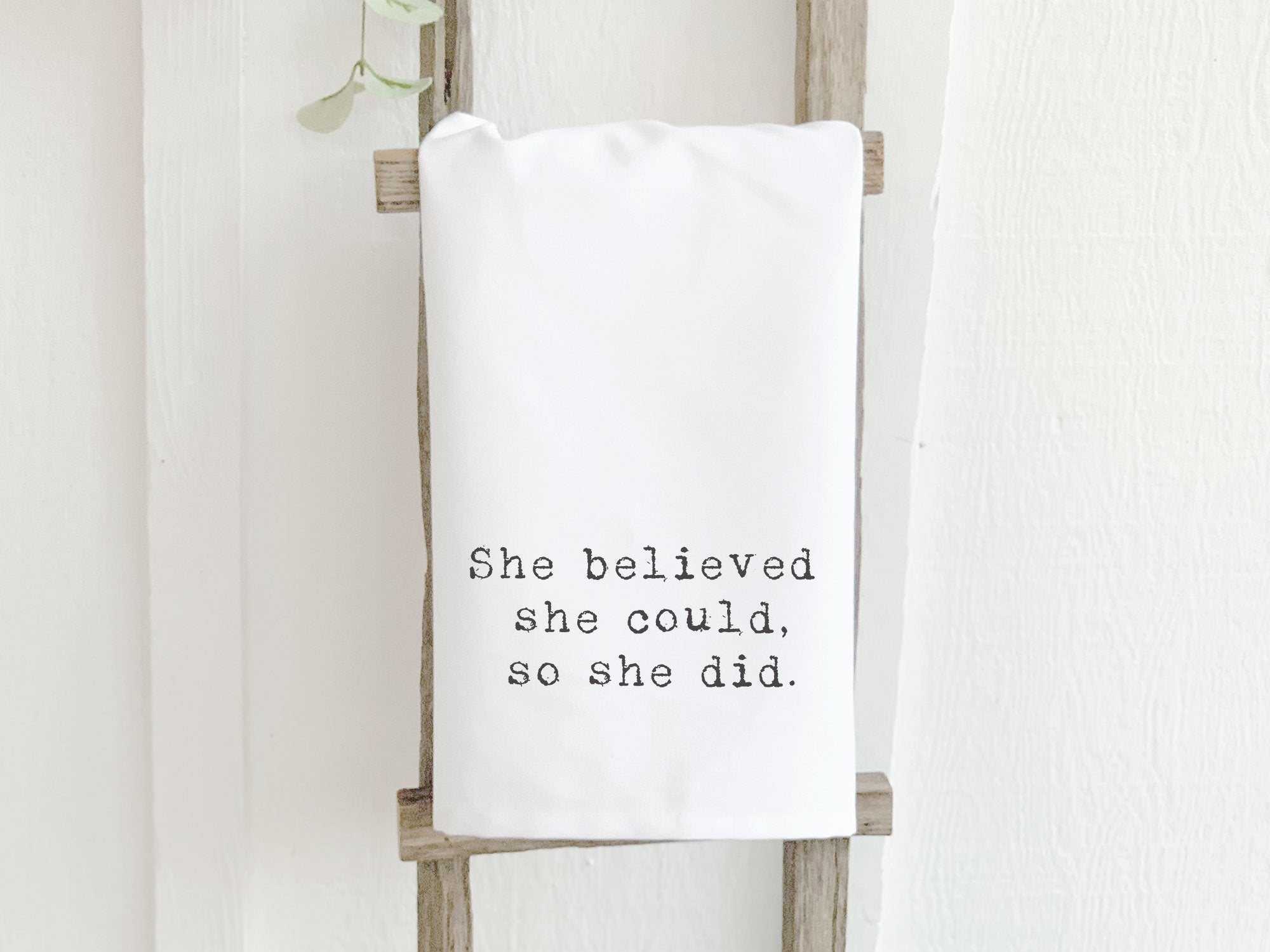 A vibrant cotton tea towel featuring the inspirational quote 'She Believed She Could, So She Did', designed with a flour sack style and hemmed edges.