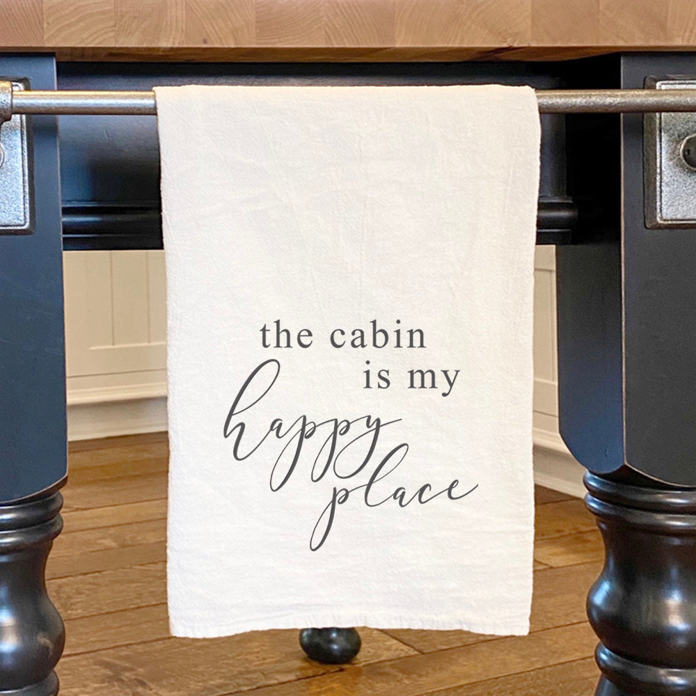 Cotton tea towel featuring 'The Cabin is My Happy Place' design, vibrant colors, and hemmed edges, perfect for kitchen use.