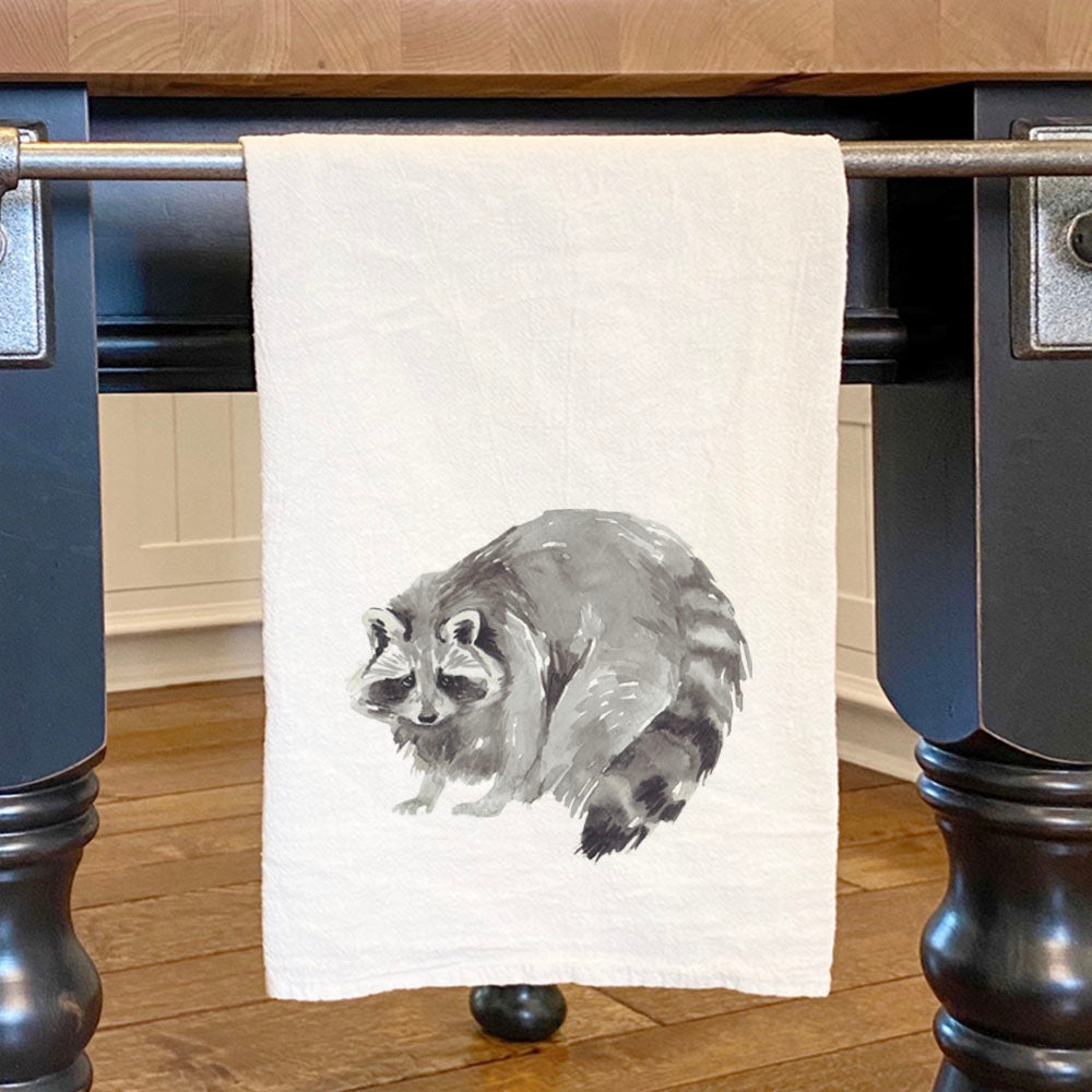 A beautifully designed cotton tea towel featuring a watercolor raccoon, showcasing vibrant colors and hemmed edges, perfect for kitchen use.