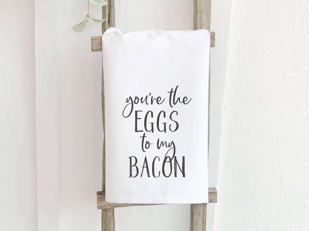 Cotton tea towel featuring the phrase 'You're the Eggs to My Bacon' with a vibrant design, perfect for kitchen use.