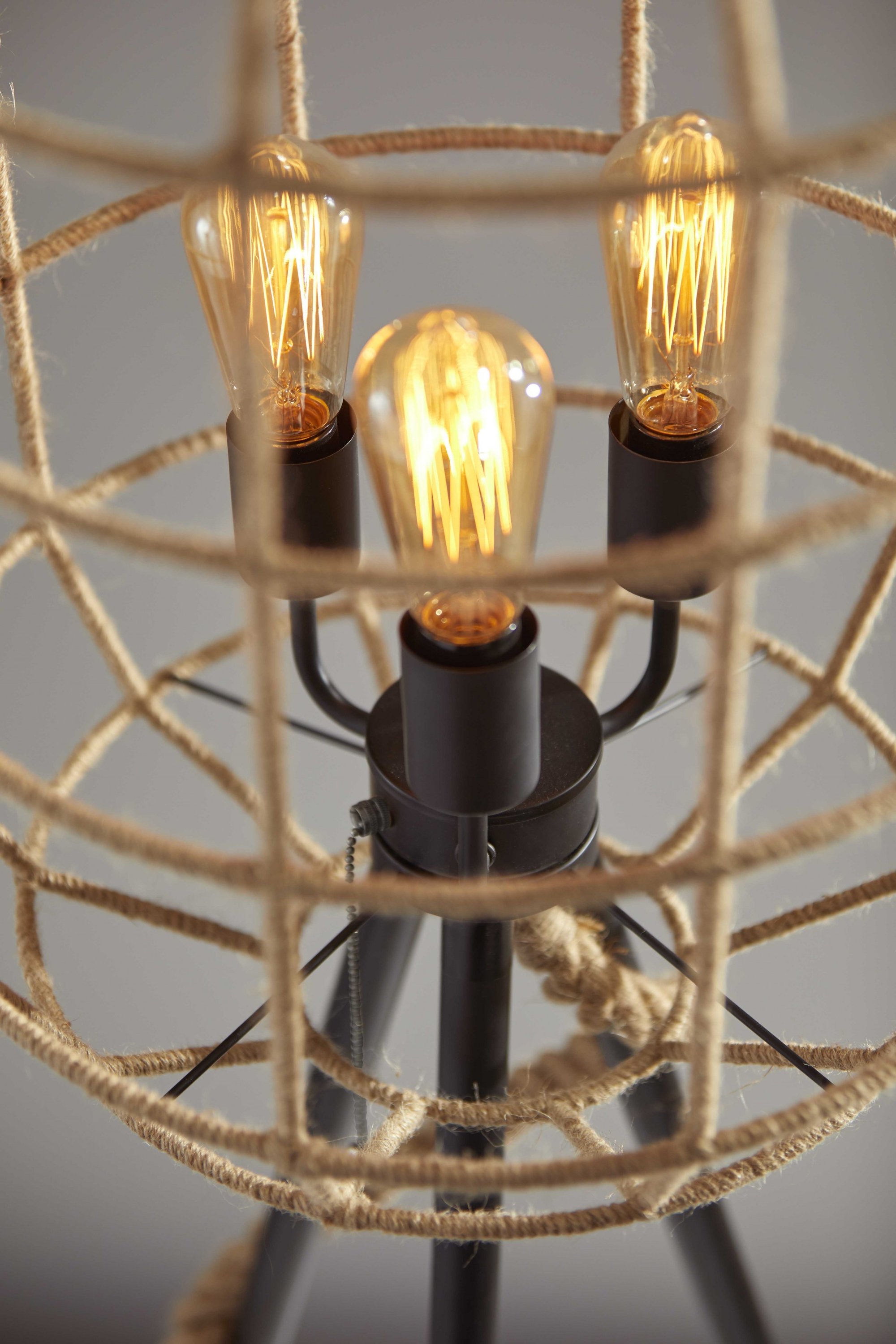 A modern black shade floor lamp with a tripod base, featuring a metal wire cage wrapped in rope and three vintage Edison bulbs.