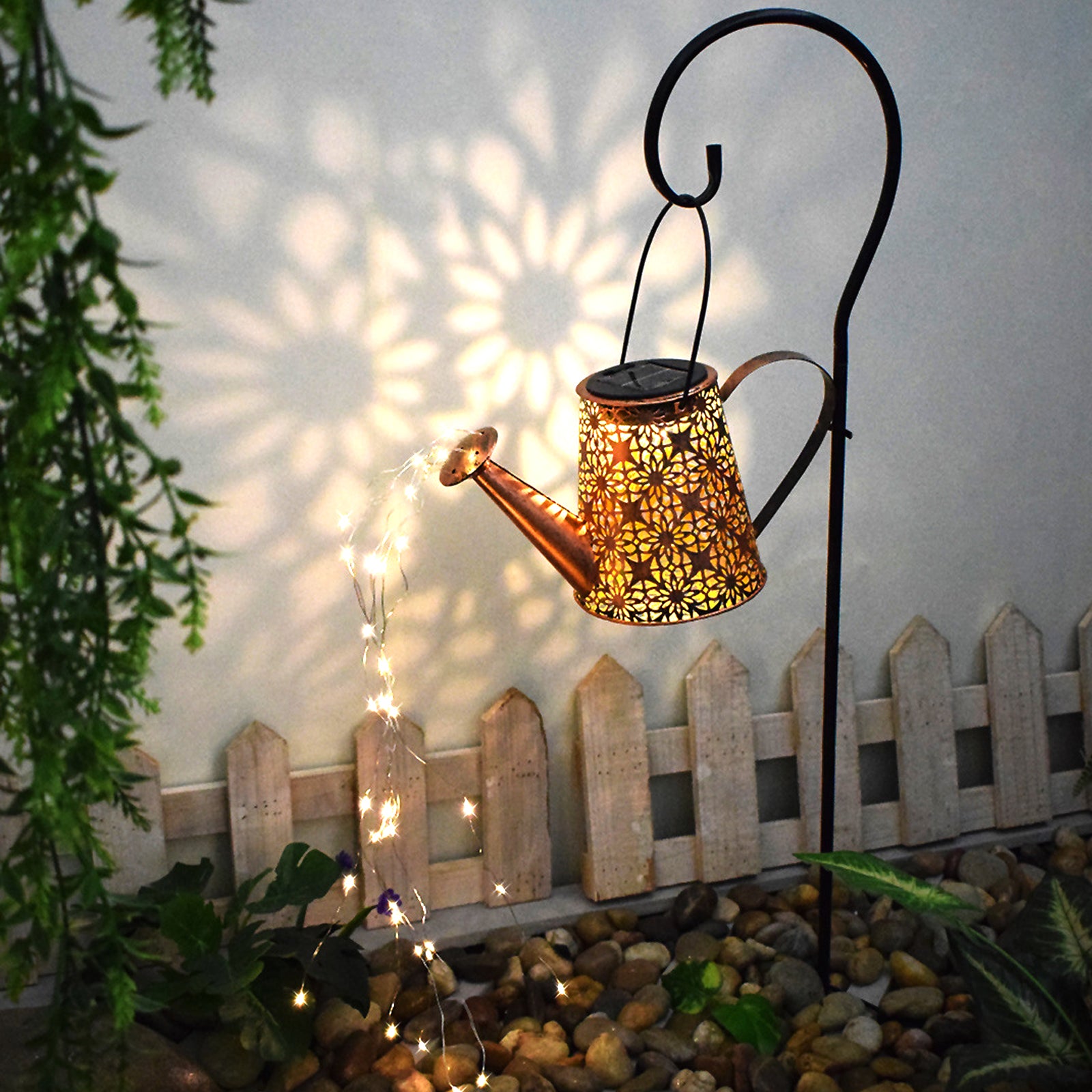 A decorative solar watering can light with LED string lights, hanging on a shepherd hook, illuminating a garden with beautiful patterns.