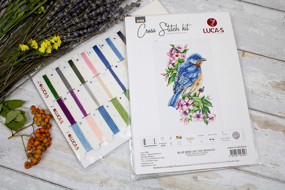 A counted cross-stitch kit featuring a blue bird on a branch, including Aida canvas, colorful threads, and a needle.