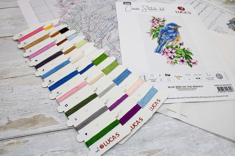 A counted cross-stitch kit featuring a blue bird on a branch, including Aida canvas, colorful threads, and a needle.