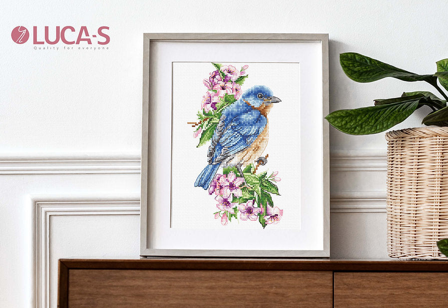 A counted cross-stitch kit featuring a blue bird on a branch, including Aida canvas, colorful threads, and a needle.
