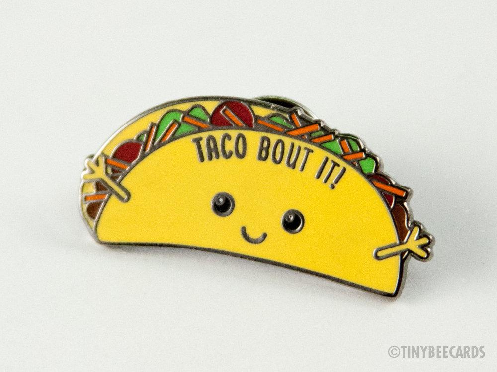 A colorful hard enamel pin featuring a kawaii taco with the pun 'Taco Bout It!' on a black nickel background.