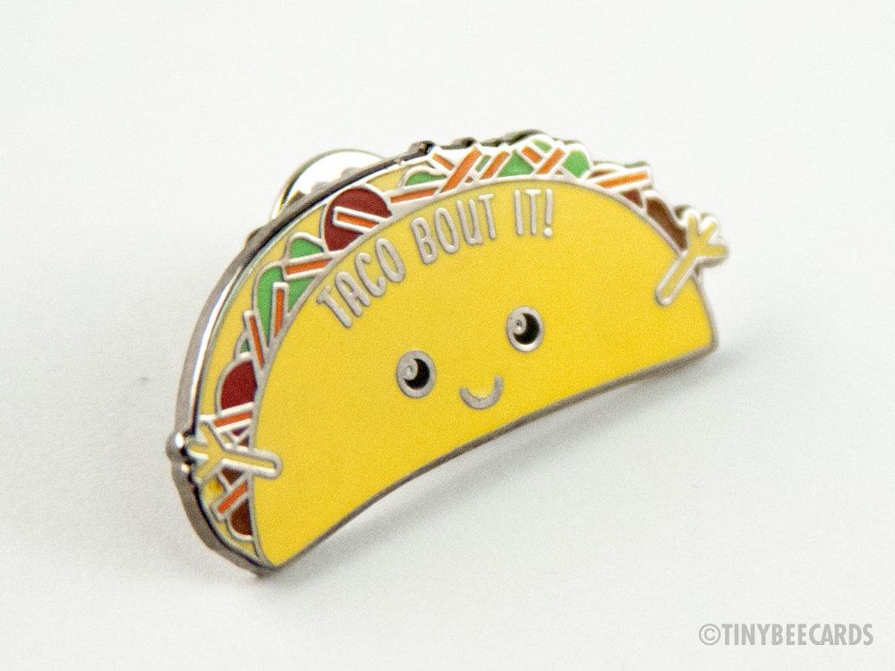 A colorful hard enamel pin featuring a kawaii taco with the pun 'Taco Bout It!' on a black nickel background.