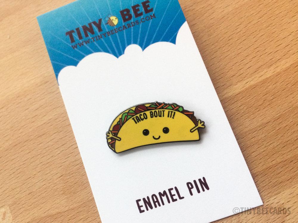 A colorful hard enamel pin featuring a kawaii taco with the pun 'Taco Bout It!' on a black nickel background.