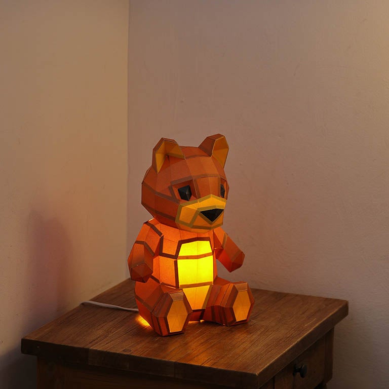 A charming 3D paper model of a teddy bear lamp, pre-cut and pre-folded, showcasing its inviting design with arms open wide.