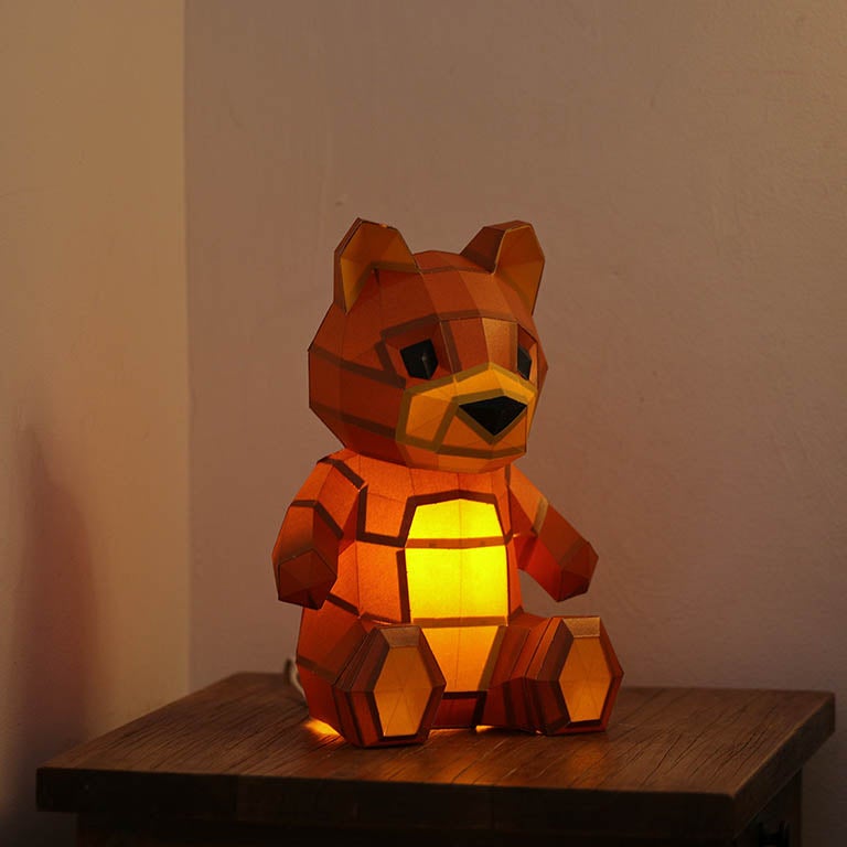 A charming 3D paper model of a teddy bear lamp, pre-cut and pre-folded, showcasing its inviting design with arms open wide.