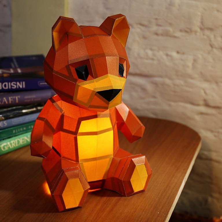 A charming 3D paper model of a teddy bear lamp, pre-cut and pre-folded, showcasing its inviting design with arms open wide.