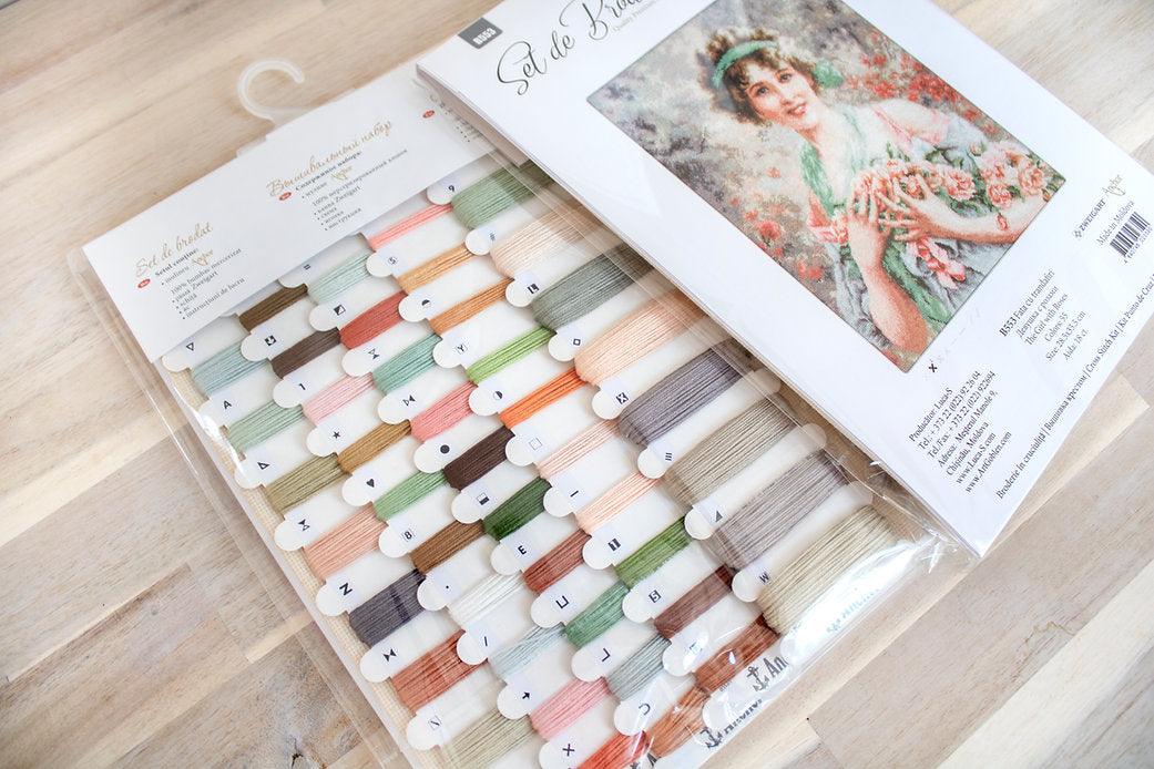 The Girl with Roses B553L Counted Cross-Stitch Kit featuring colorful threads, Aida canvas, and a needle.