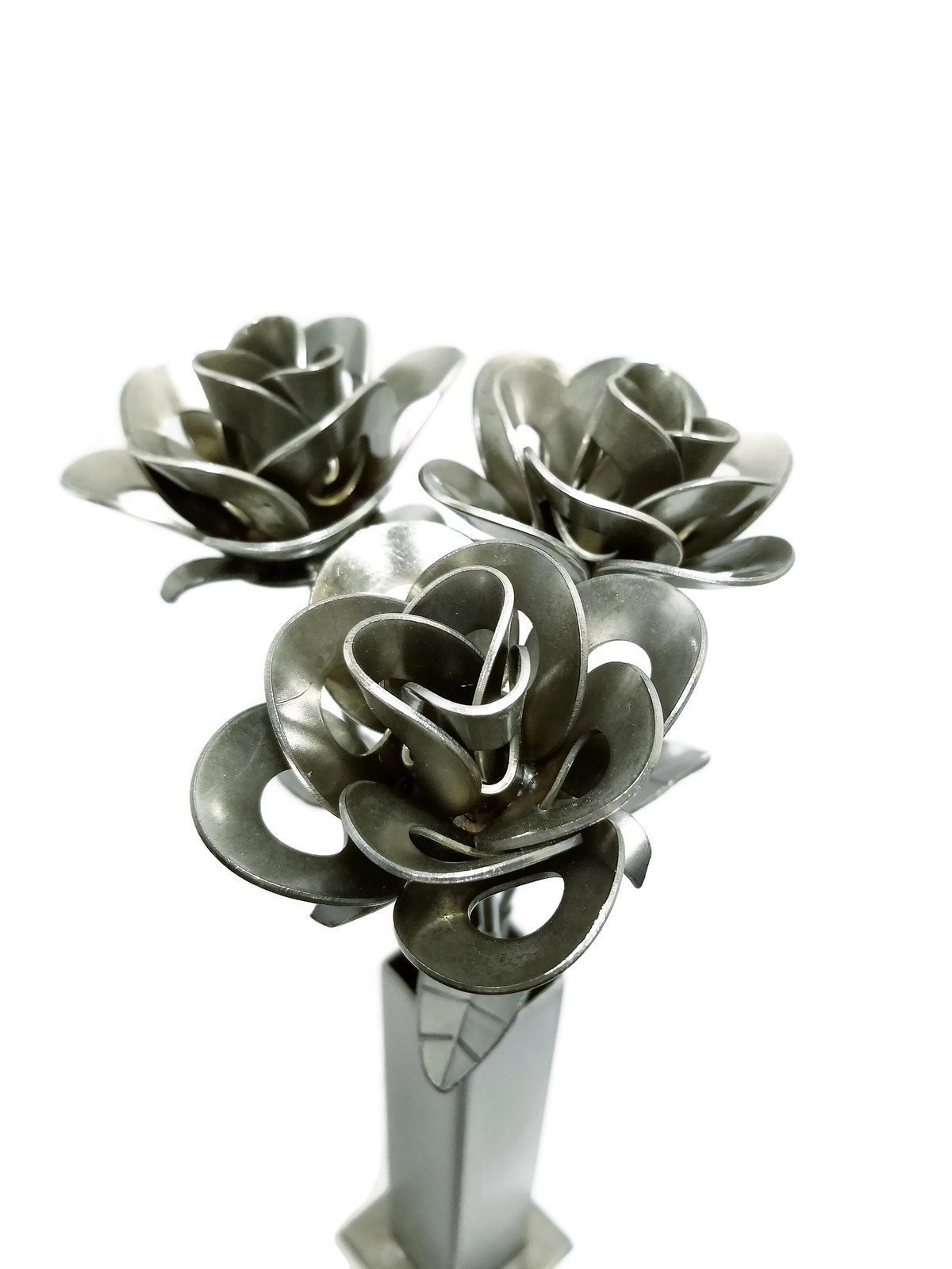 Three handcrafted metal roses in a rustic vase, made from recycled materials, showcasing unique welded designs and a shiny lacquer finish.