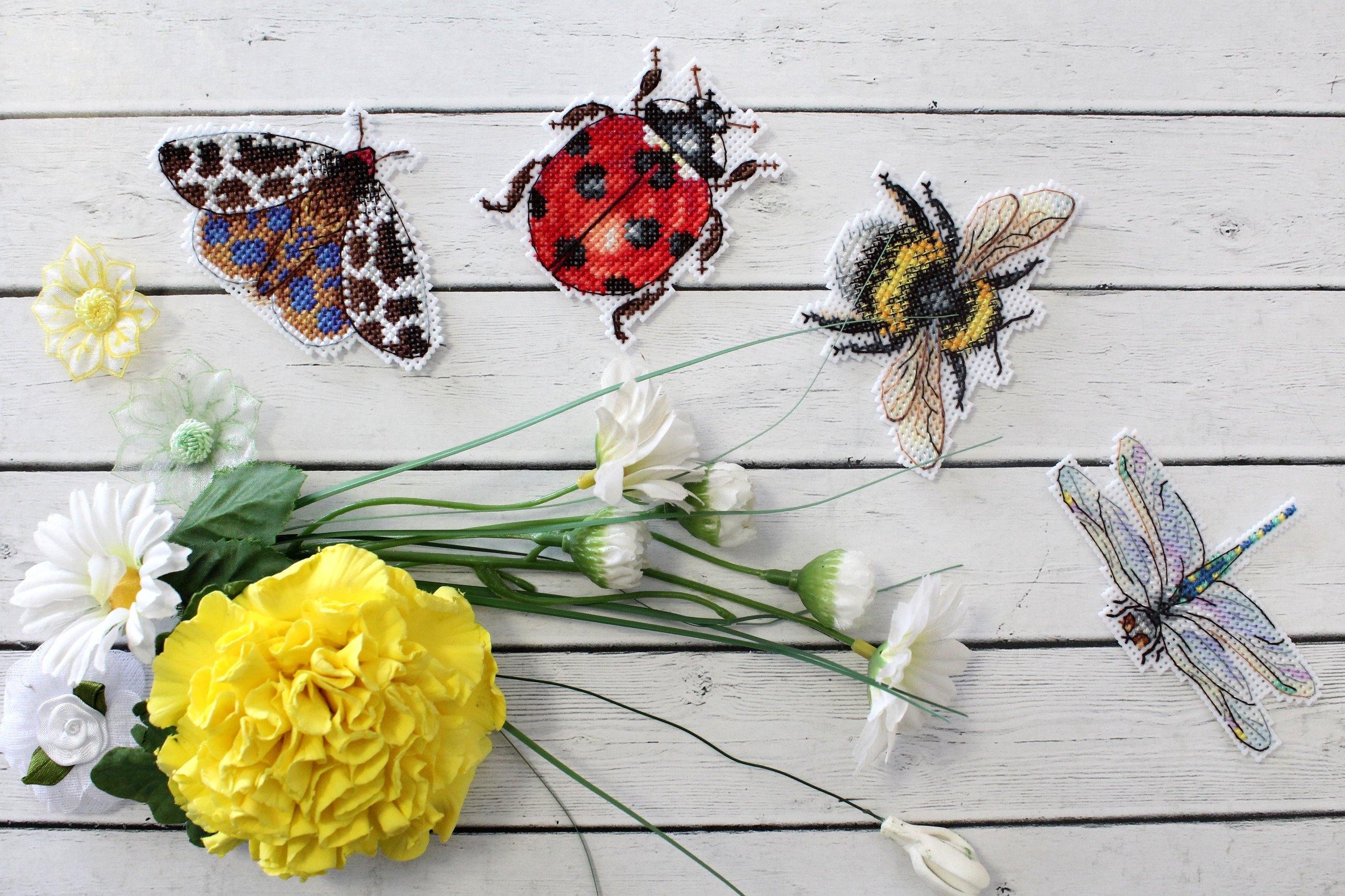A set of four colorful insect-themed cross-stitch magnets featuring a bee and ladybug, crafted from plastic canvas and cotton threads.