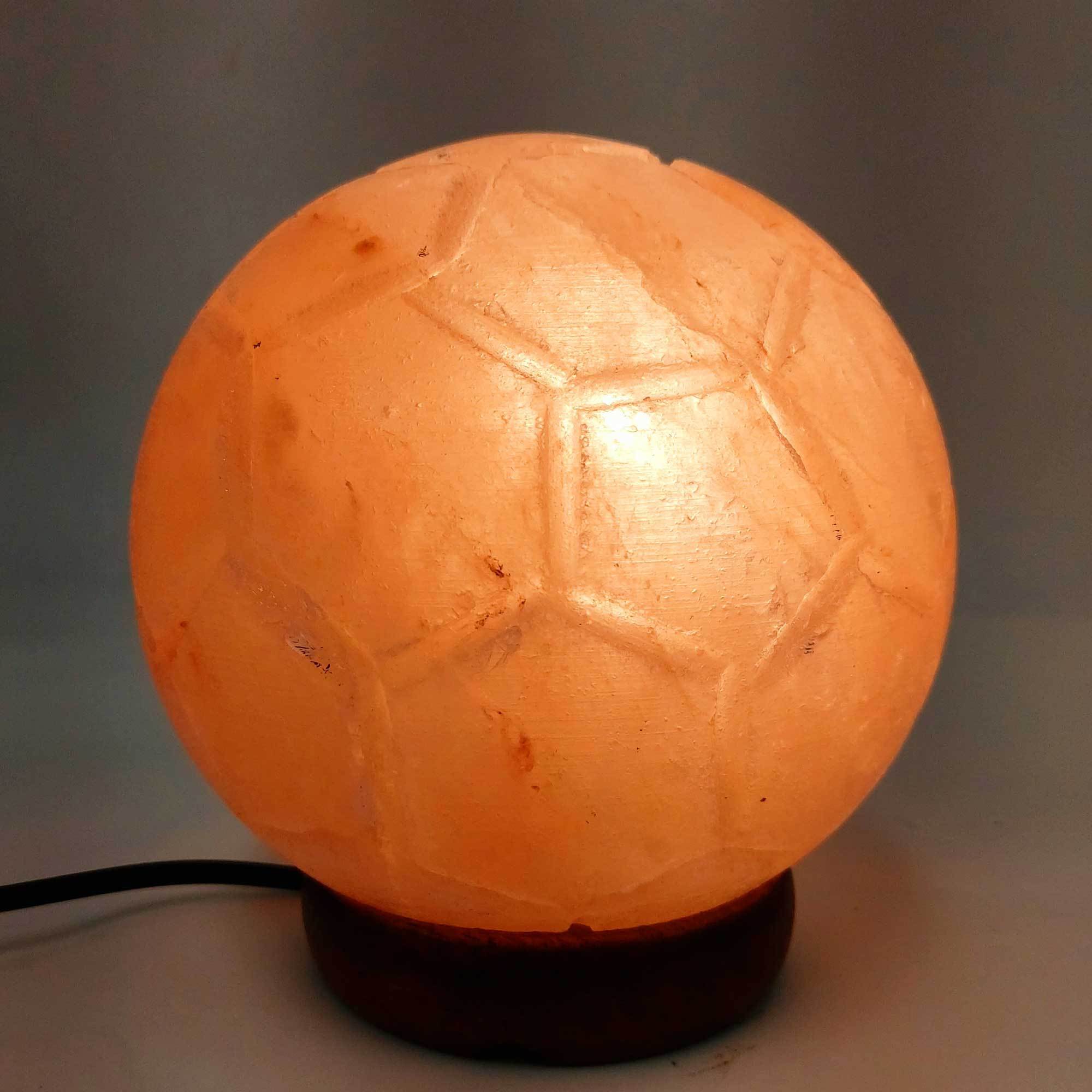 A beautifully carved Himalayan pink salt lamp in the shape of a soccer ball, glowing softly on a wooden base.