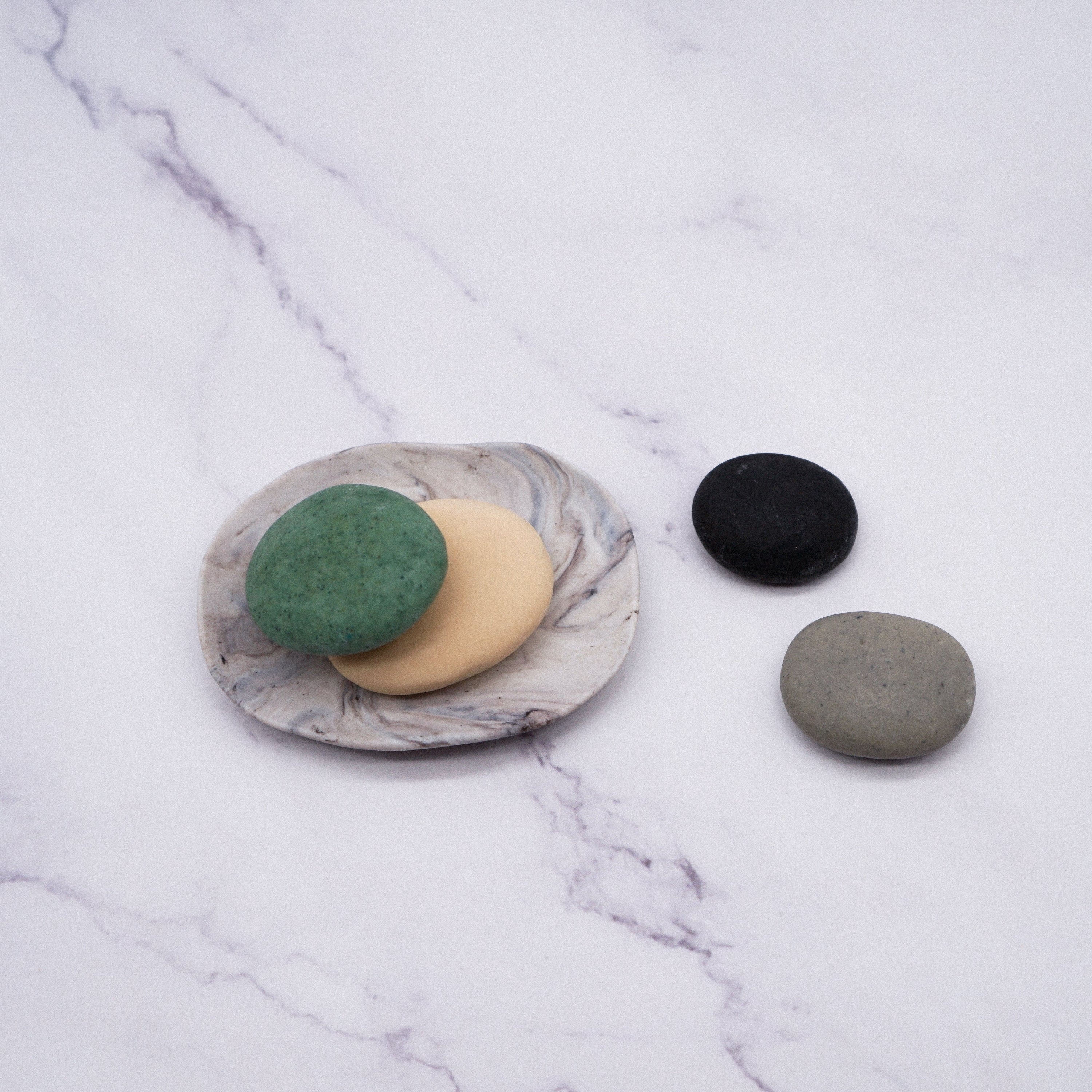 A set of assorted handmade big pebble soaps in a white porcelain dish with a marble effect design, showcasing their natural beauty.