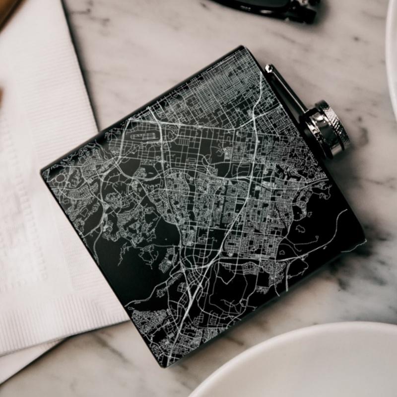 Custom engraved matte black hip flask featuring a map of Irvine, California with coordinates, accompanied by a canvas bag and funnel.