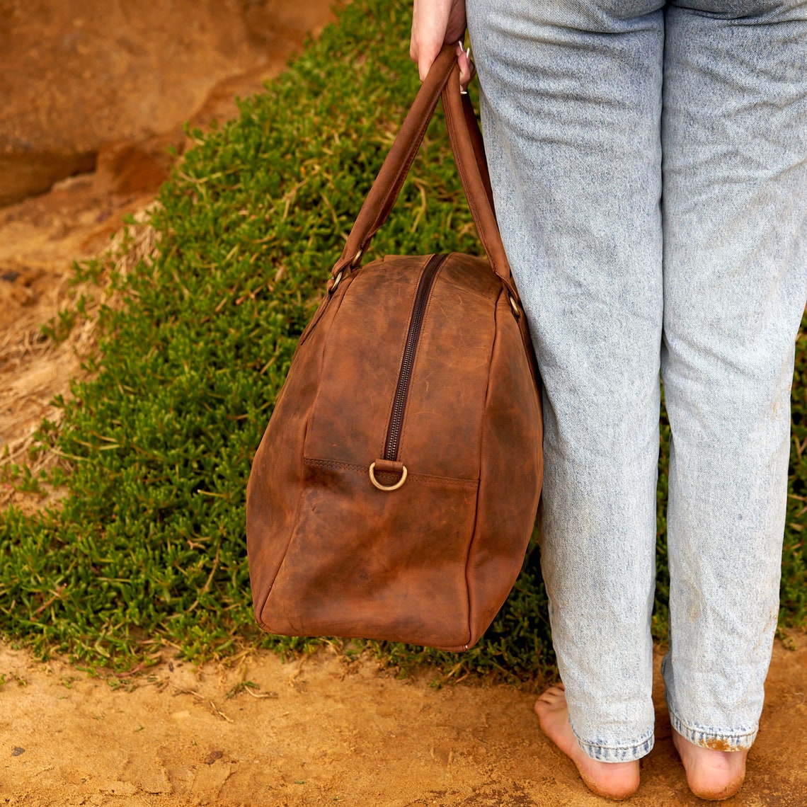 Handmade rustic vintage leather duffle bag with brass zippers, adjustable shoulder strap, and spacious interior, perfect for weekend getaways.