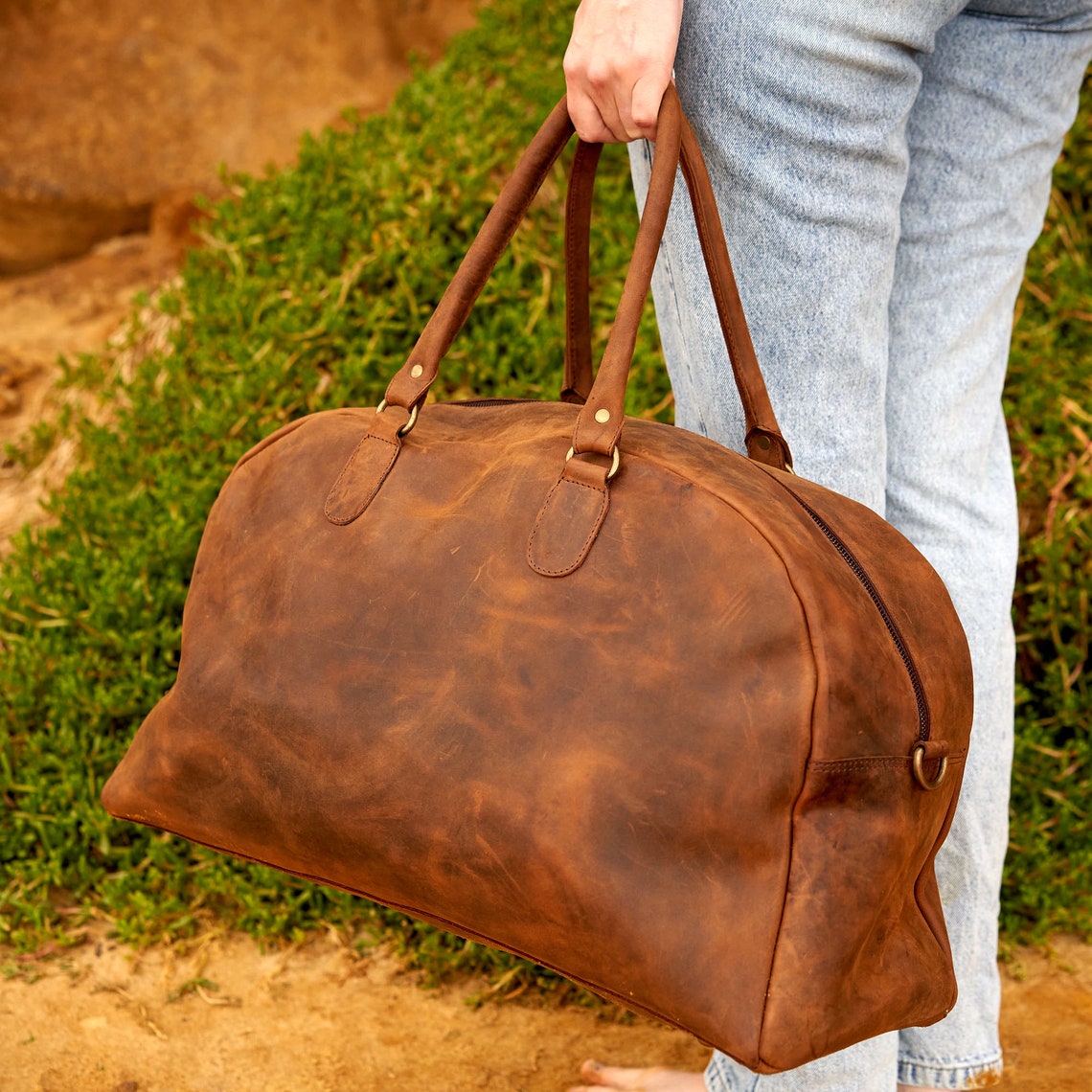Handmade rustic vintage leather duffle bag with brass zippers, adjustable shoulder strap, and spacious interior, perfect for weekend getaways.