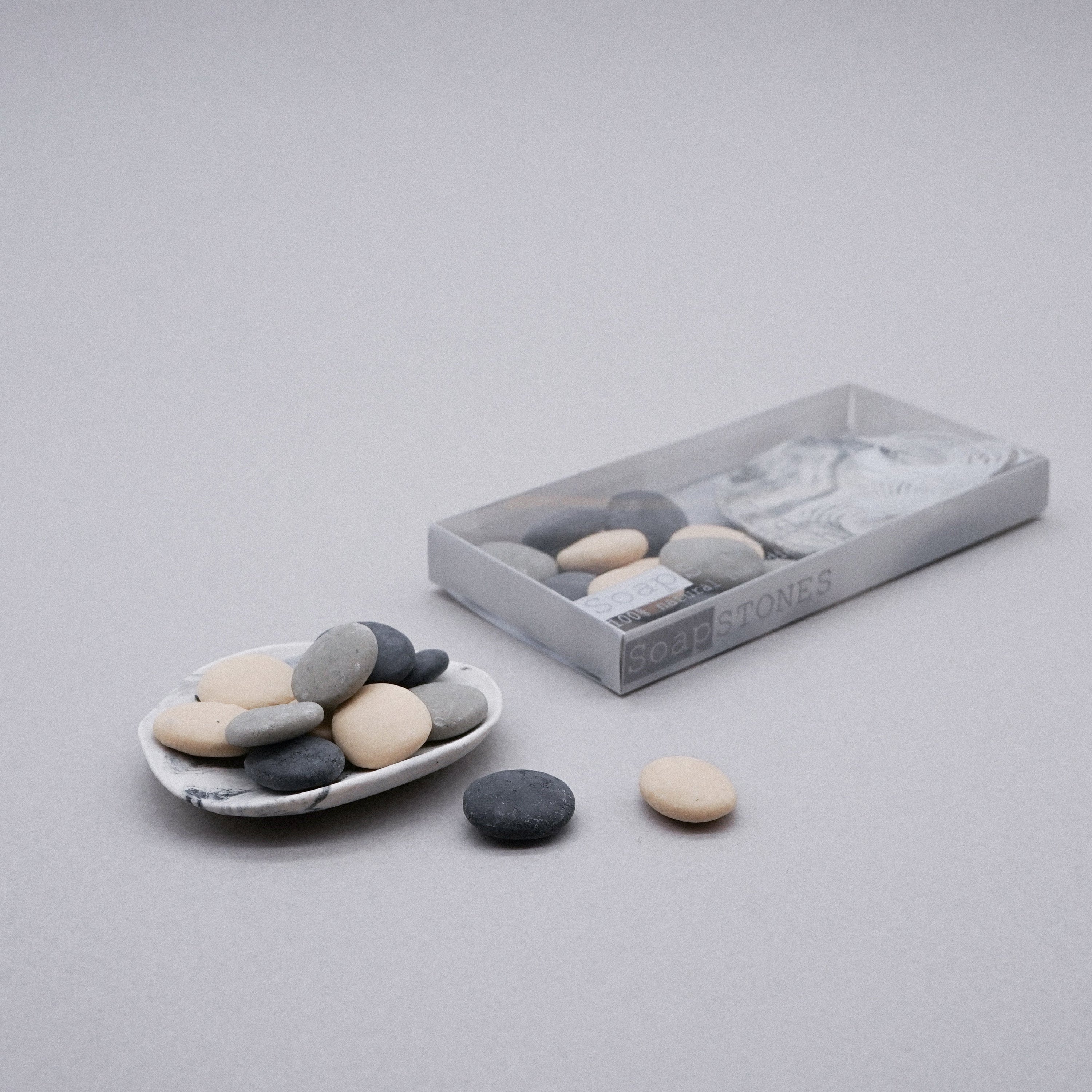 A set of small pebble-shaped handmade soaps in a white porcelain dish with a grey marble effect, showcasing their natural design.