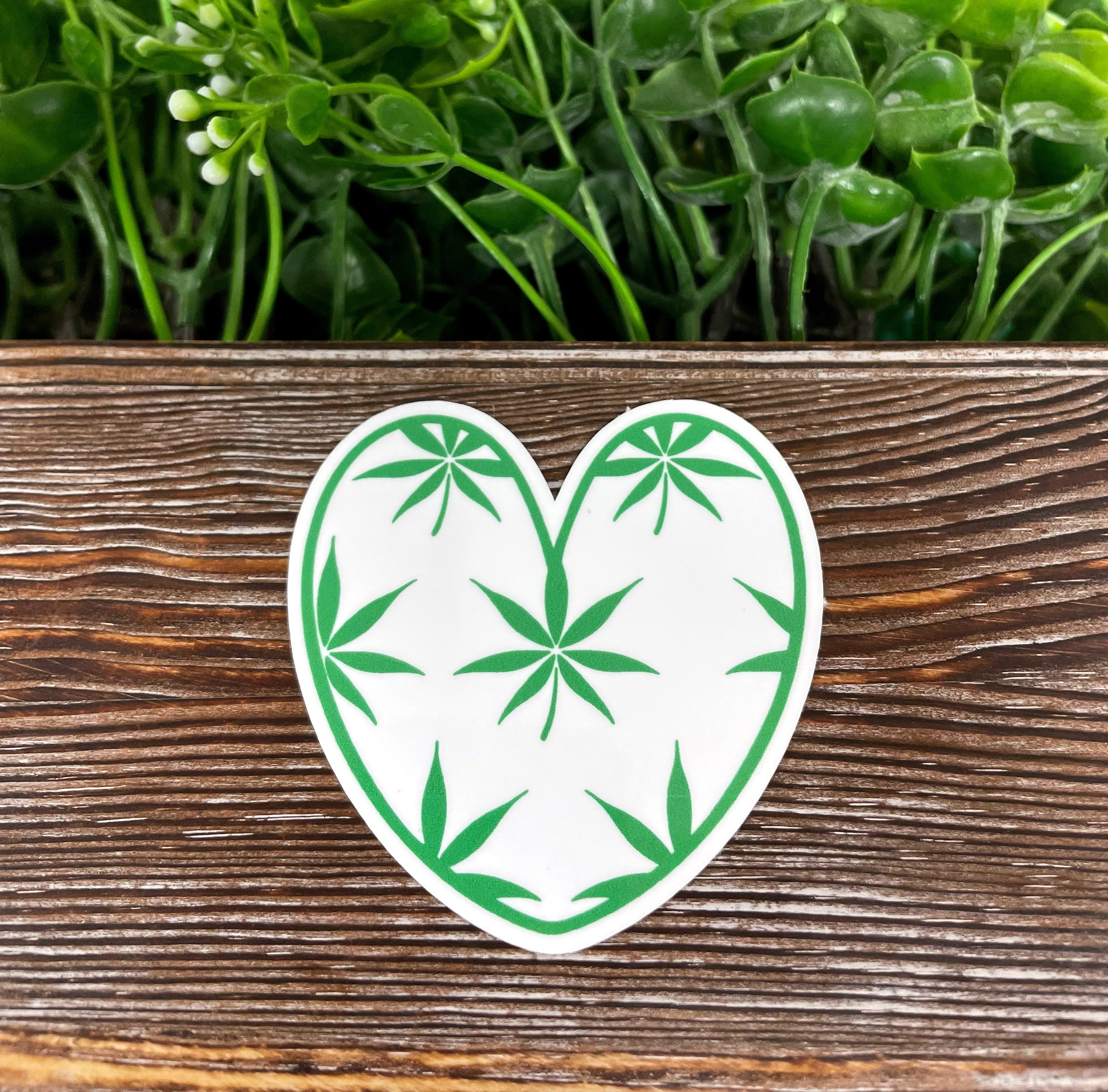 A vibrant Cannabis Leaf Heart die cut vinyl sticker on a smooth surface, showcasing its intricate design and water-resistant laminate.