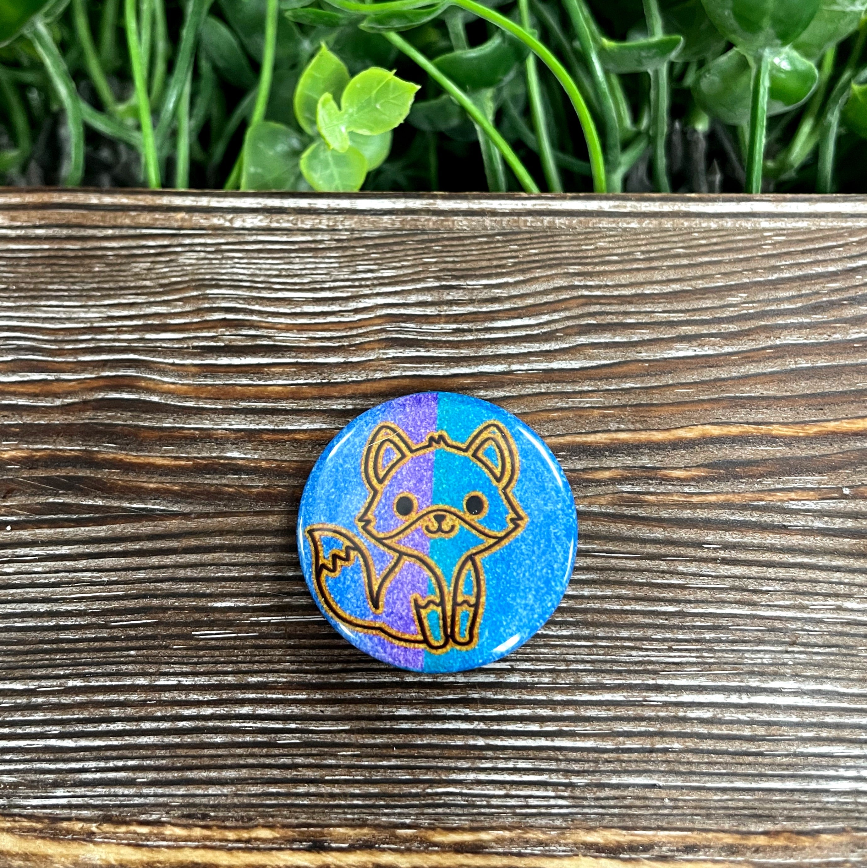 A 1.25-inch Fox Outline Graphic Art Button featuring blue and purple glitter, showcasing a unique design perfect for personal accessories.