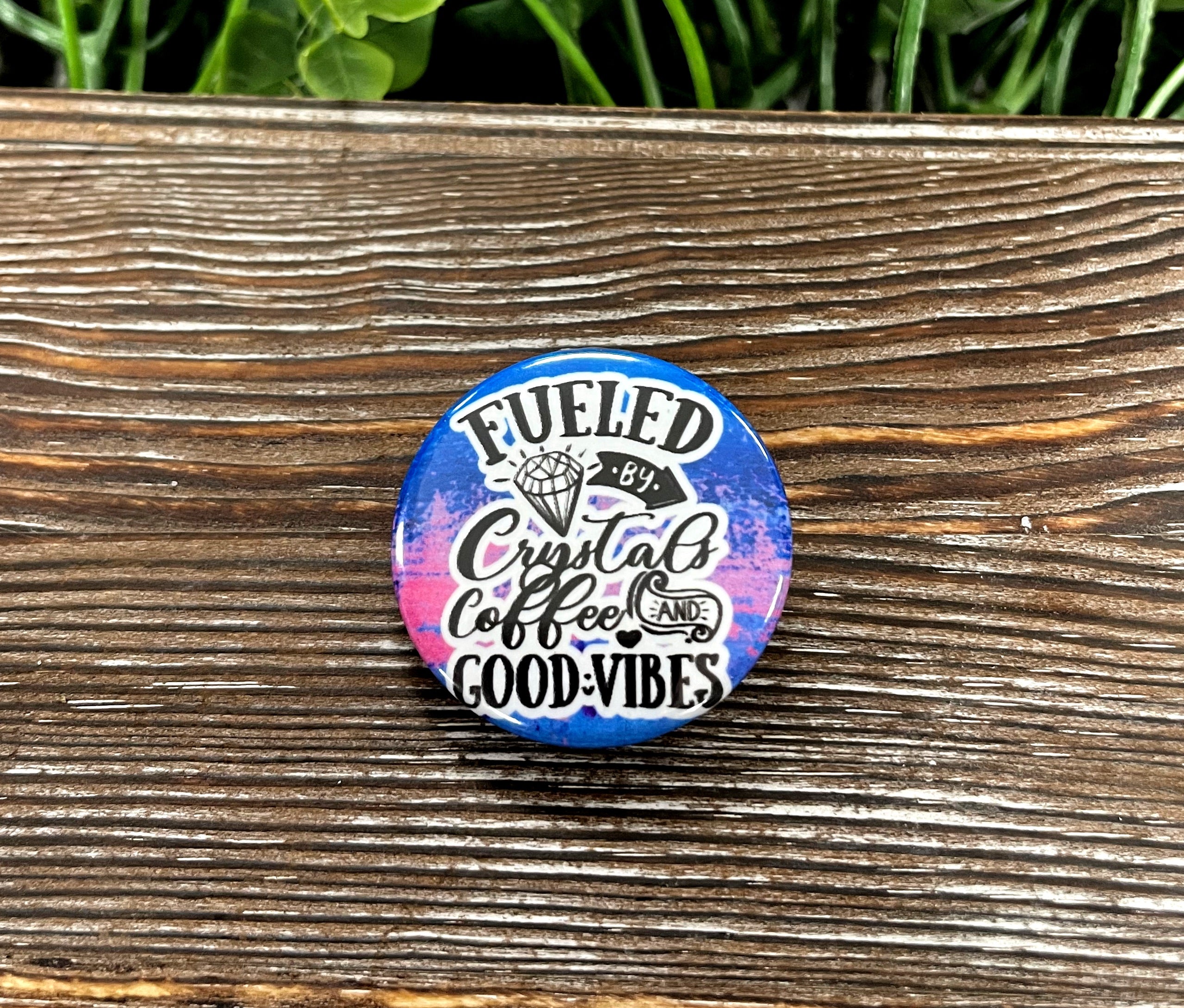 A vibrant blue and pink graphic art button featuring the phrase 'Fueled by Crystals and Caffeine', perfect for personalizing accessories.