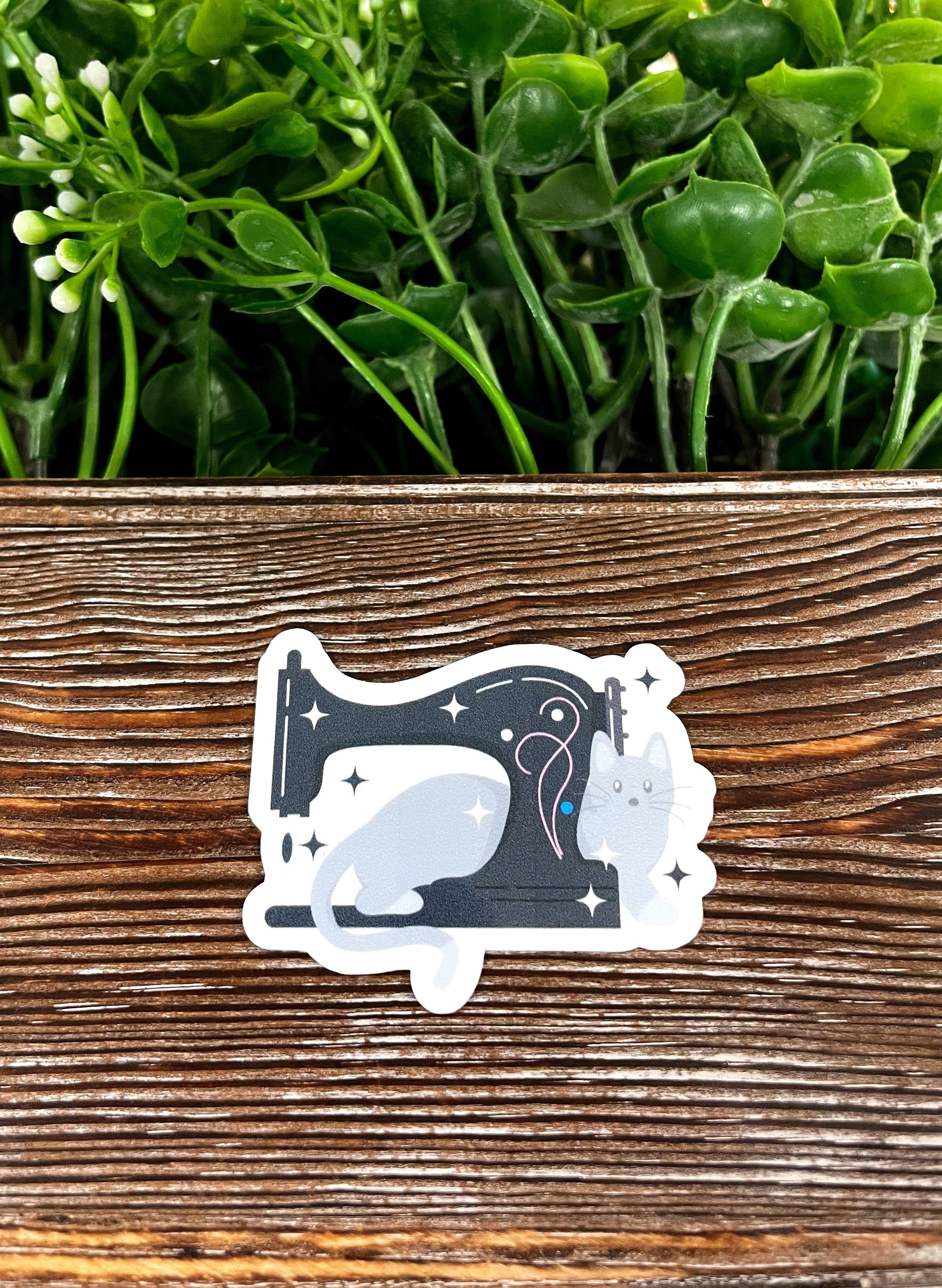Gray cat sitting beside a black sewing machine, designed as a die cut vinyl sticker with a boho aesthetic.