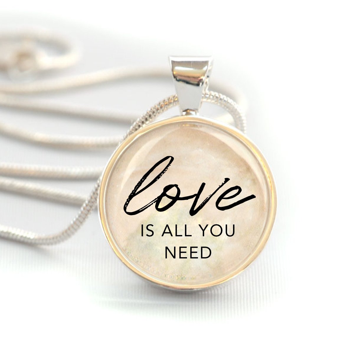 A silver-plated pendant necklace featuring the phrase 'Love Is All You Need' under a glass charm, strung on a 20-inch silver-plated chain.