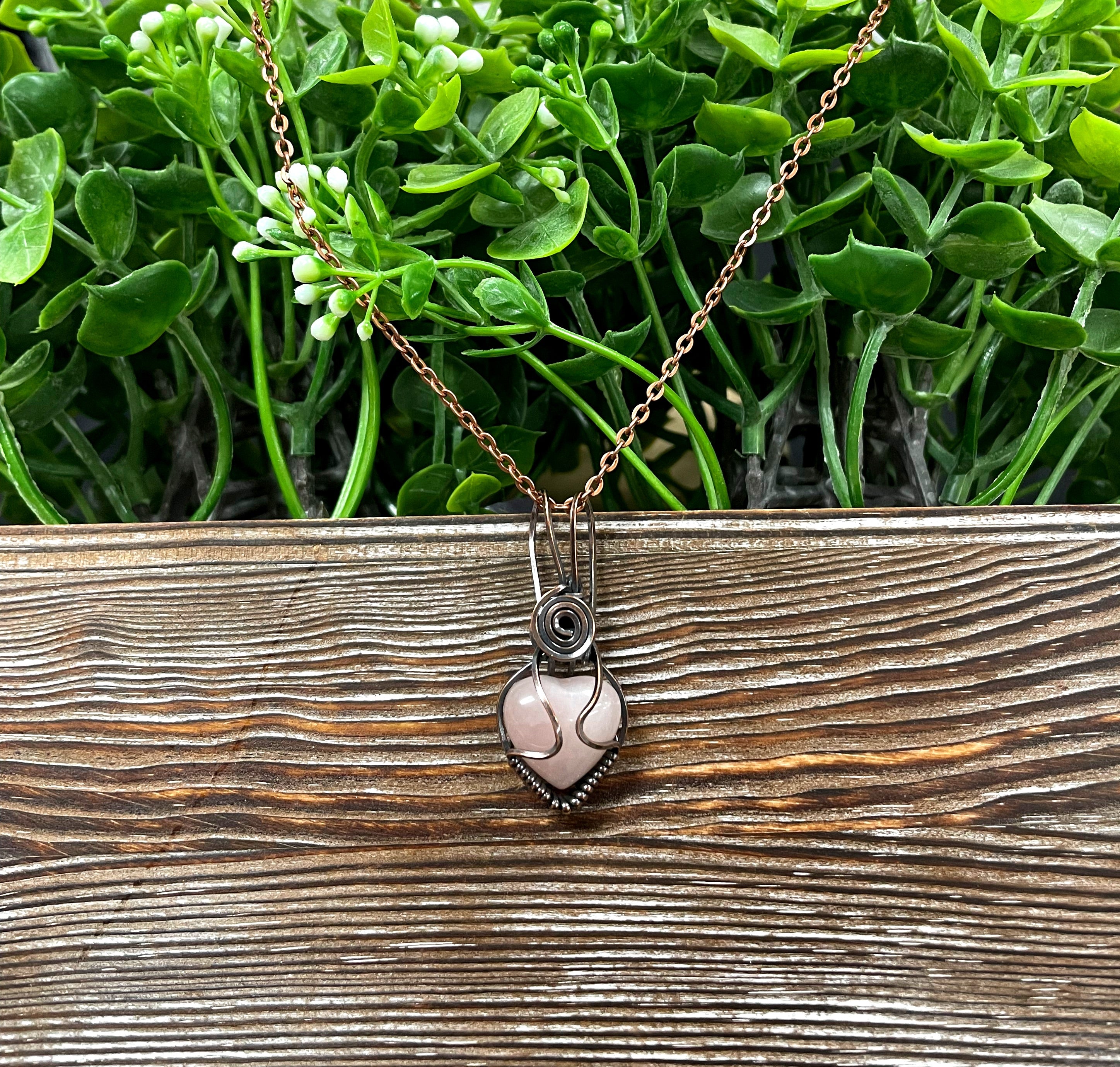 Small heart-shaped pendant made of pink rose quartz gemstone, wrapped in oxidized copper wire, displayed on a 21-inch cord.