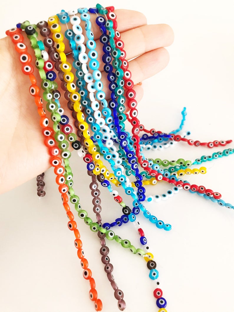A collection of 6mm to 12mm flat round Evil Eye Glass Beads in various colors, displayed in a hand for size reference.