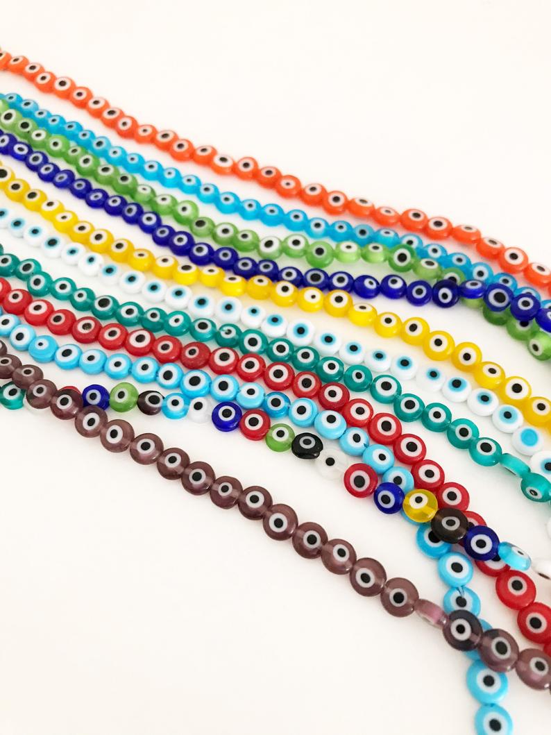 A collection of 6mm to 12mm flat round Evil Eye Glass Beads in various colors, displayed in a hand for size reference.