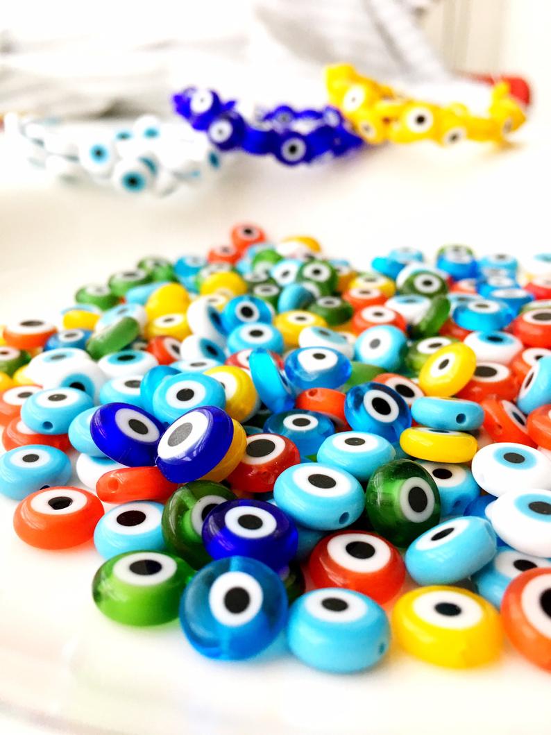 A collection of 6mm to 12mm flat round Evil Eye Glass Beads in various colors, displayed in a hand for size reference.