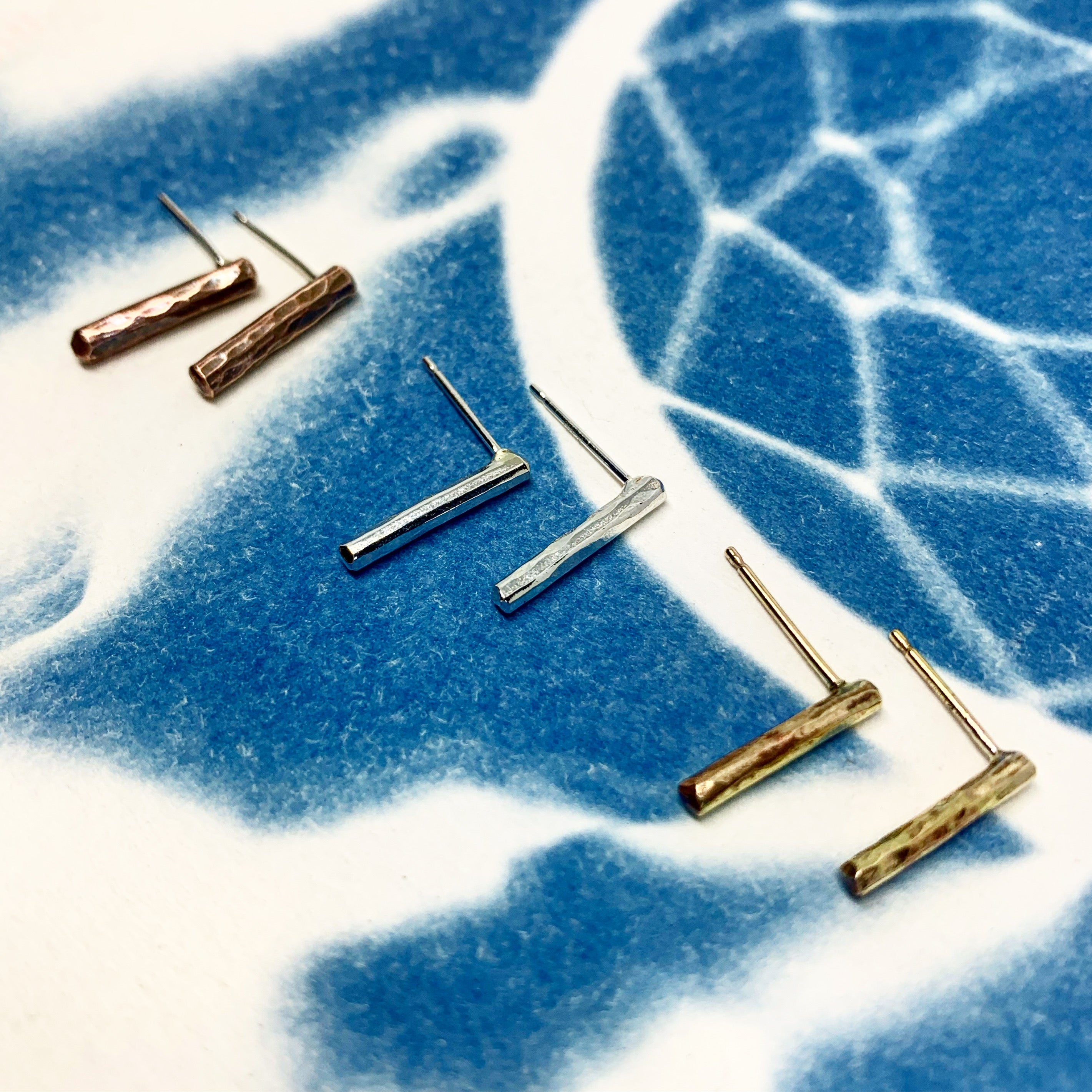 A pair of Balance Stud Earrings - 1/2" featuring tiny hammered rods in copper, sterling silver, and yellow gold fill, showcasing their minimalist design.