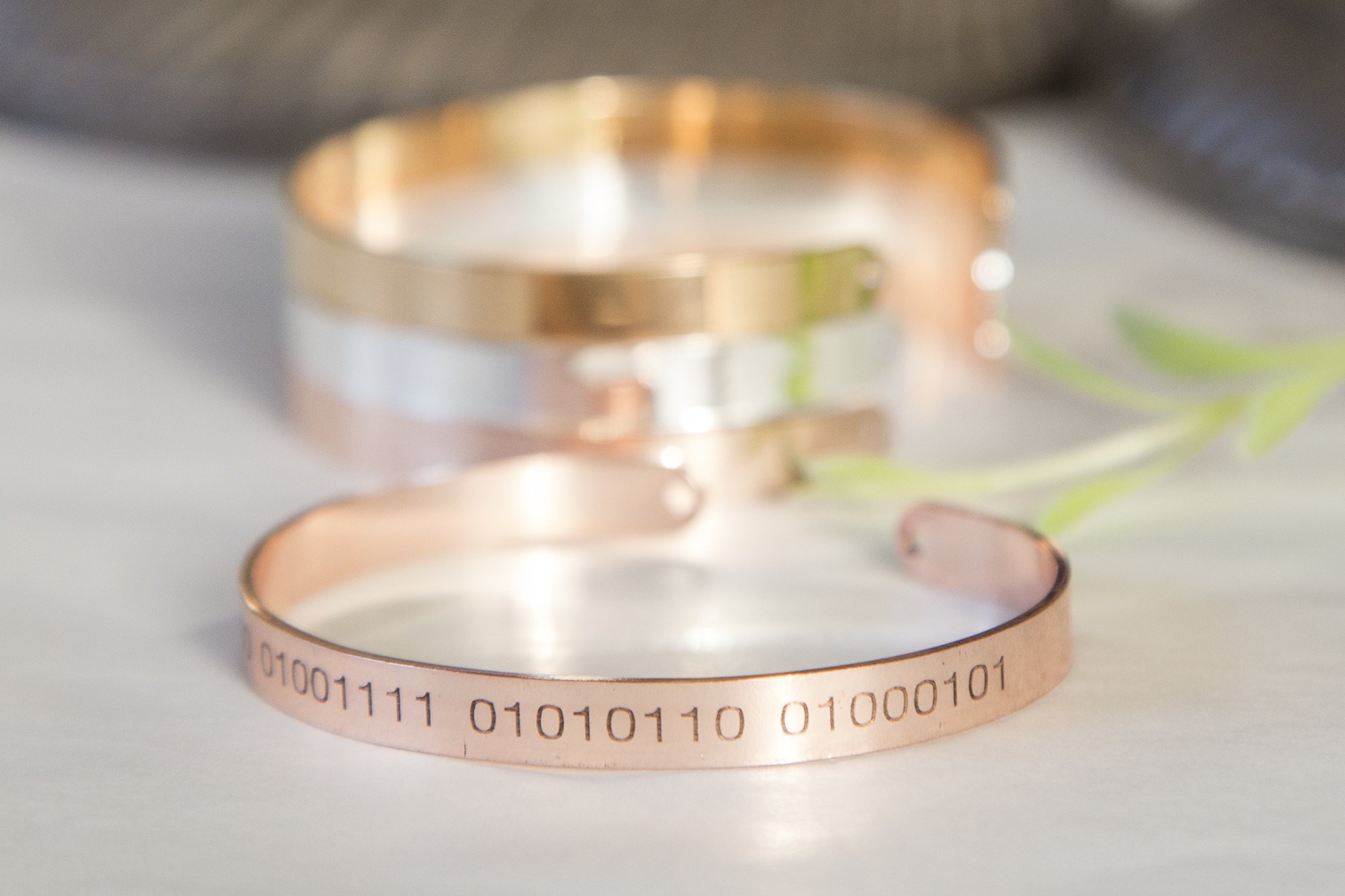 A stylish Binary Code Bracelet featuring custom engraved binary code on a delicate cuff, available in gold, rose gold, and silver plating.