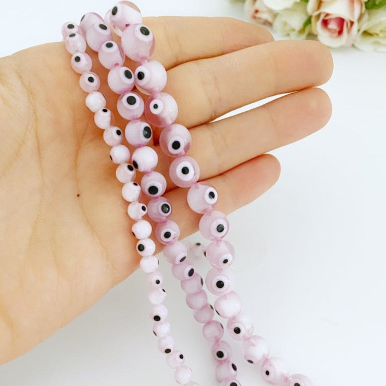 A collection of pale pink evil eye beads in various sizes (6mm, 8mm, 10mm) displayed on a hand, showcasing their round shape and glass material.
