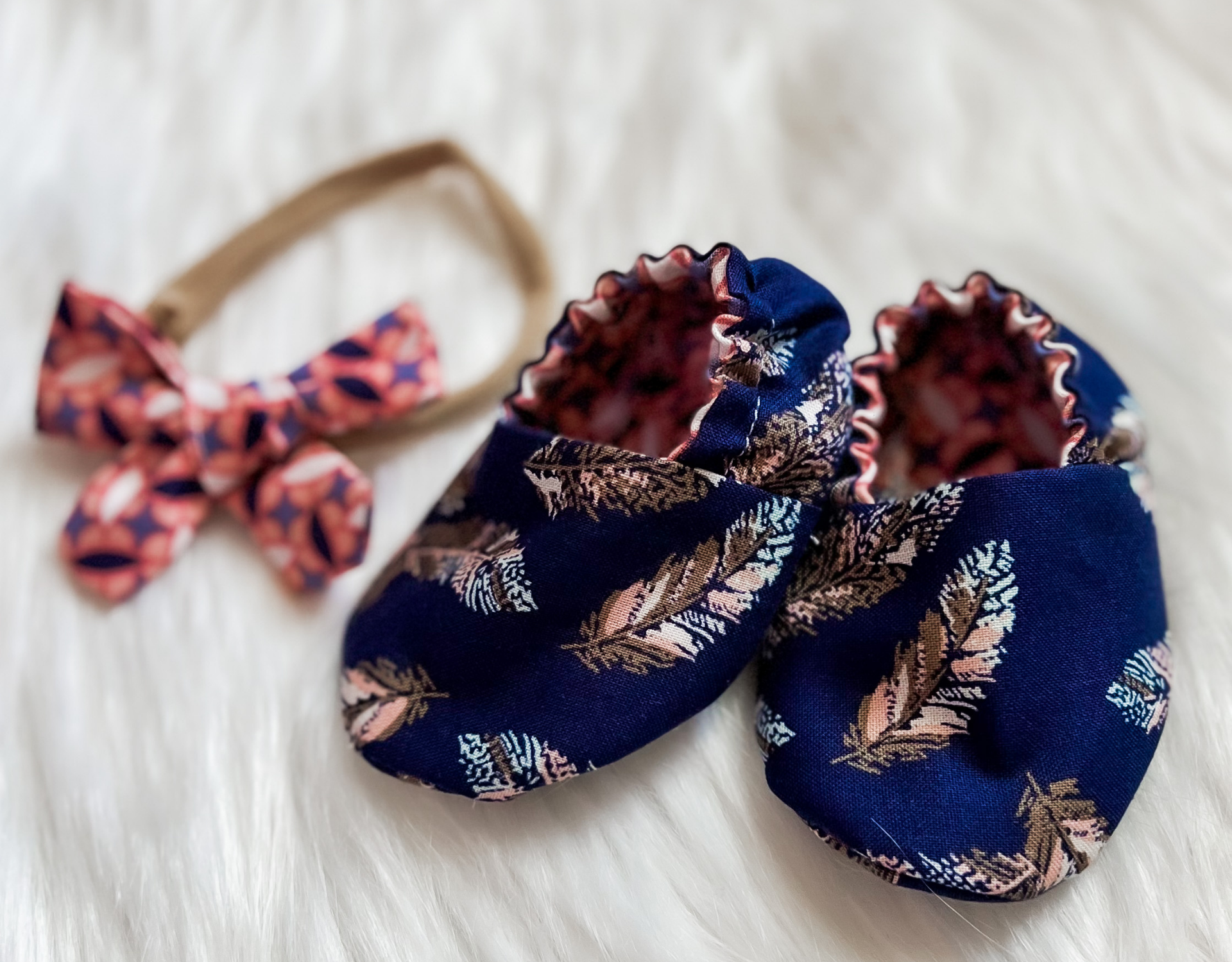 Navy blue crib shoes with feather pattern and matching bow set on a tan headband, perfect for babies.