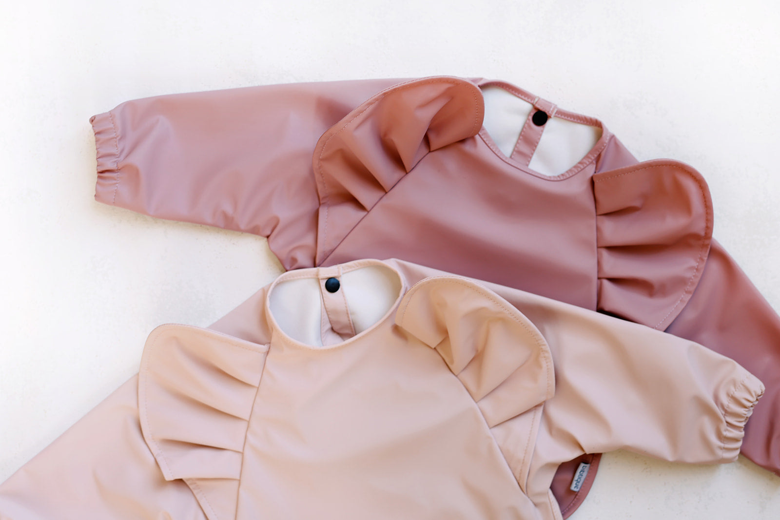 Peony Long Sleeve Ruffle Angel Bib in soft pink with ruffles, designed for babies aged 6-12 months, featuring a button closure and built-in pocket.