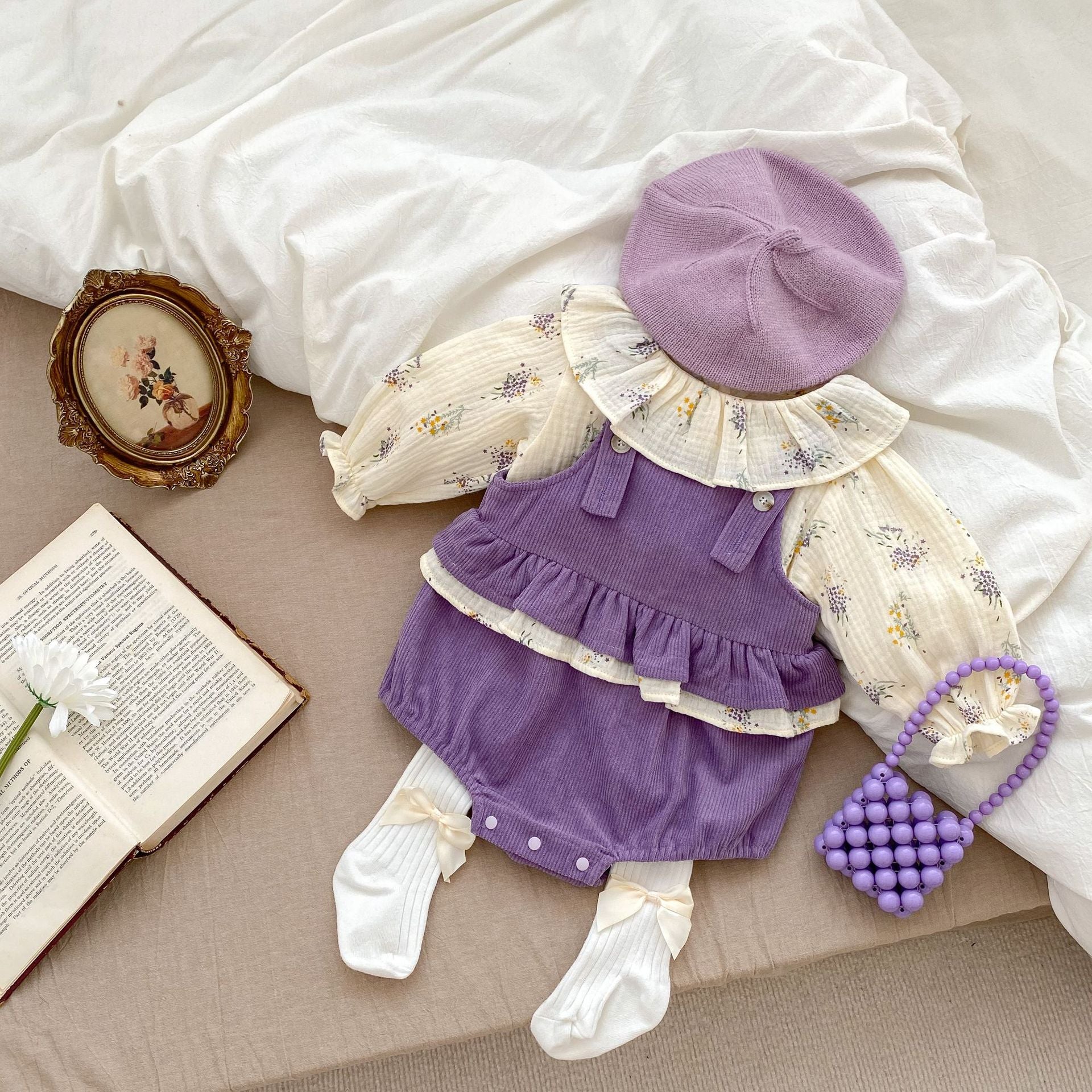 A cute baby girl wearing a long-sleeved floral top with purple straps, showcasing a vibrant floral pattern.