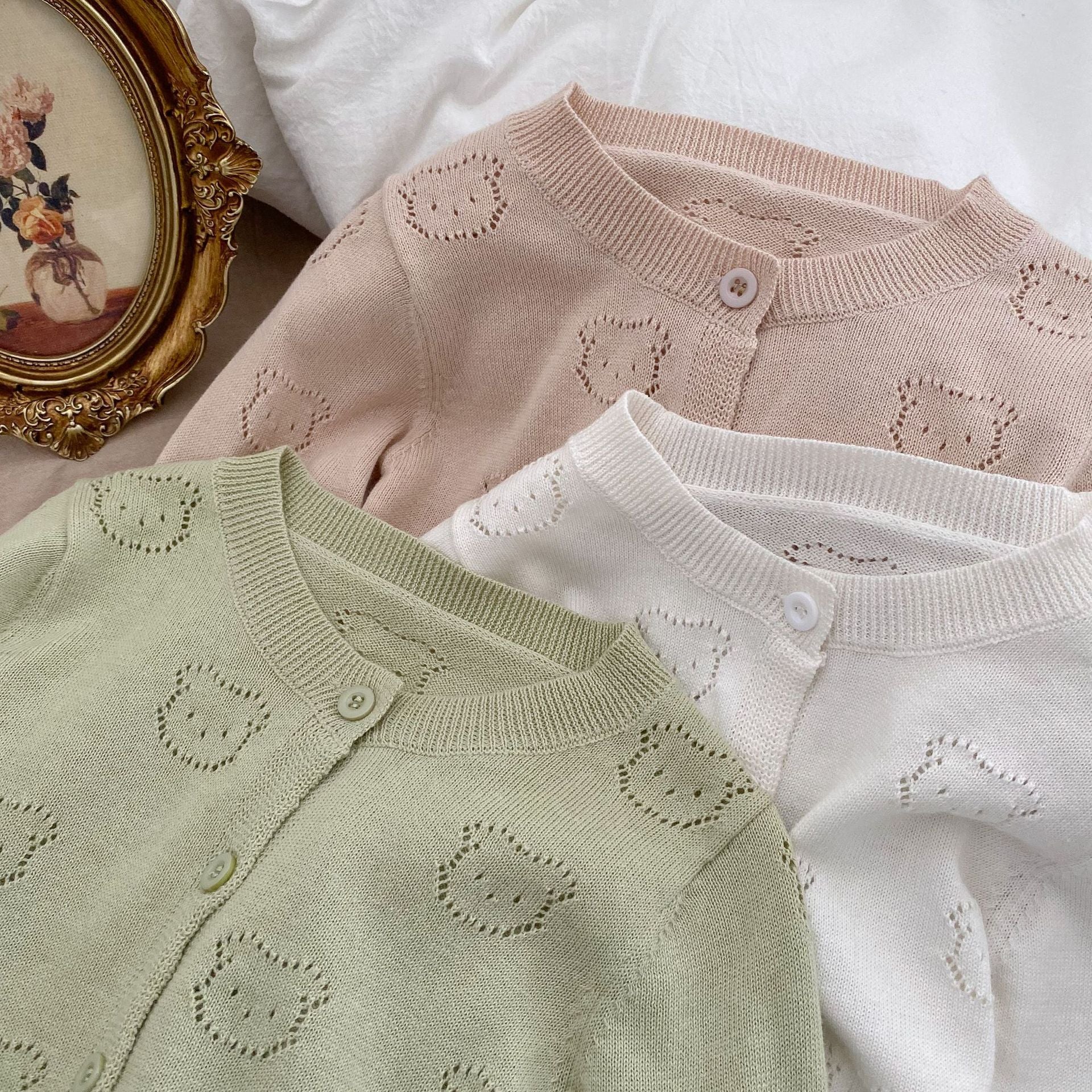 A soft cotton cardigan for baby girls featuring a hollow carved design, available in white, pink, and green.