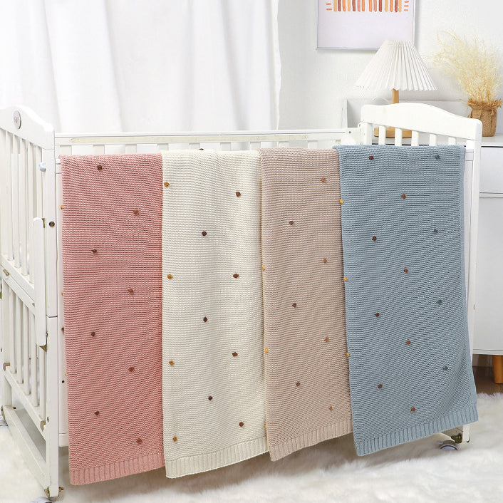 A cozy knitted solid color baby blanket featuring hand-embroidered pom poms, available in white, pink, blue, and khaki.