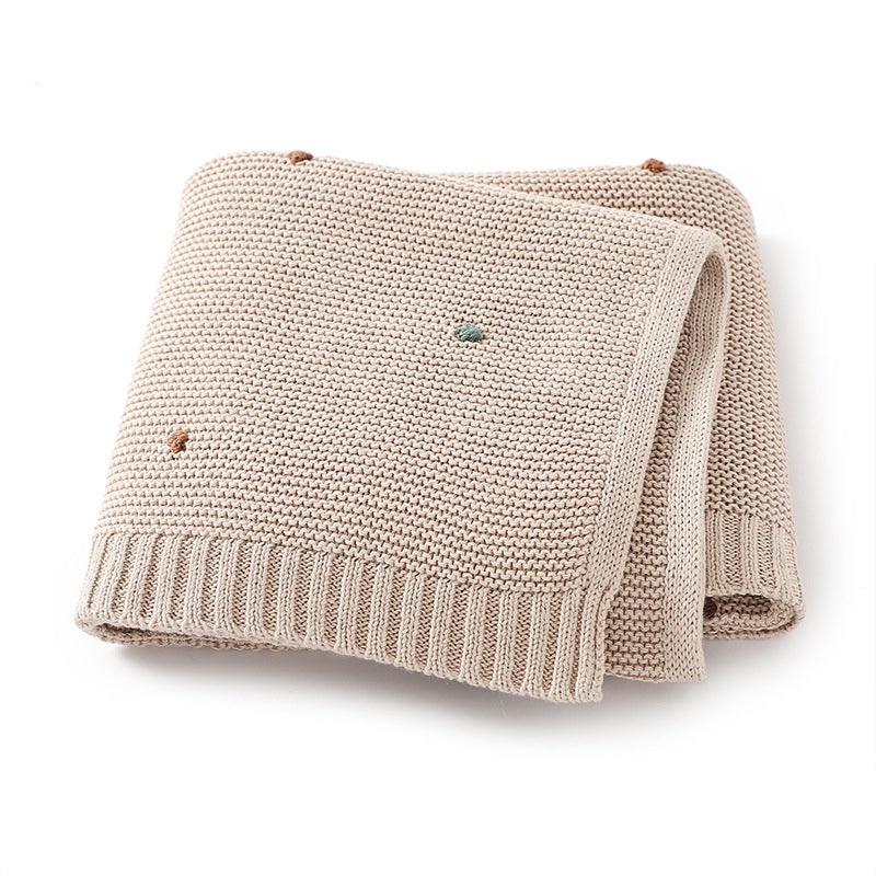 A cozy knitted solid color baby blanket featuring hand-embroidered pom poms, available in white, pink, blue, and khaki.
